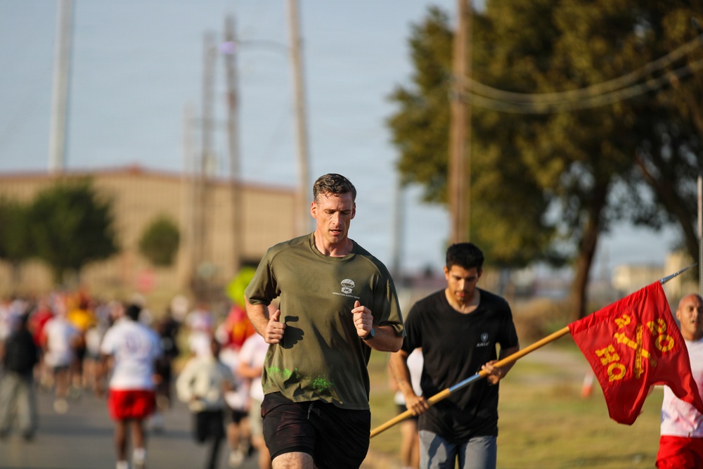 A group of runners participating in what appears to be a military or organized road race event. The lead runner, a middle-aged man in an olive green t-shirt and black shorts, is prominently in focus. A red flag with yellow lettering, consistent with a Marine Corps unit guidon, is visible on the right side of the frame.