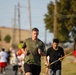 A group of runners participating in what appears to be a military or organized road race event. The lead runner, a middle-aged man in an olive green t-shirt and black shorts, is prominently in focus. A red flag with yellow lettering, consistent with a Marine Corps unit guidon, is visible on the right side of the frame.