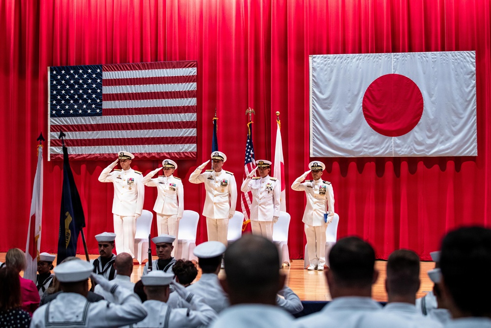 A formal military ceremony is taking place on a stage decorated with red curtains, featuring both the United States flag and the Japanese flag displayed prominently. Five officers in white dress uniforms are standing at salute on the stage, while enlisted personnel in white uniforms observe from the audience. The setting suggests a joint US-Japan naval ceremony, likely a change of command or official military function.