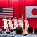 A formal military ceremony is taking place on a stage decorated with red curtains, featuring both the United States flag and the Japanese flag displayed prominently. Five officers in white dress uniforms are standing at salute on the stage, while enlisted personnel in white uniforms observe from the audience. The setting suggests a joint US-Japan naval ceremony, likely a change of command or official military function.