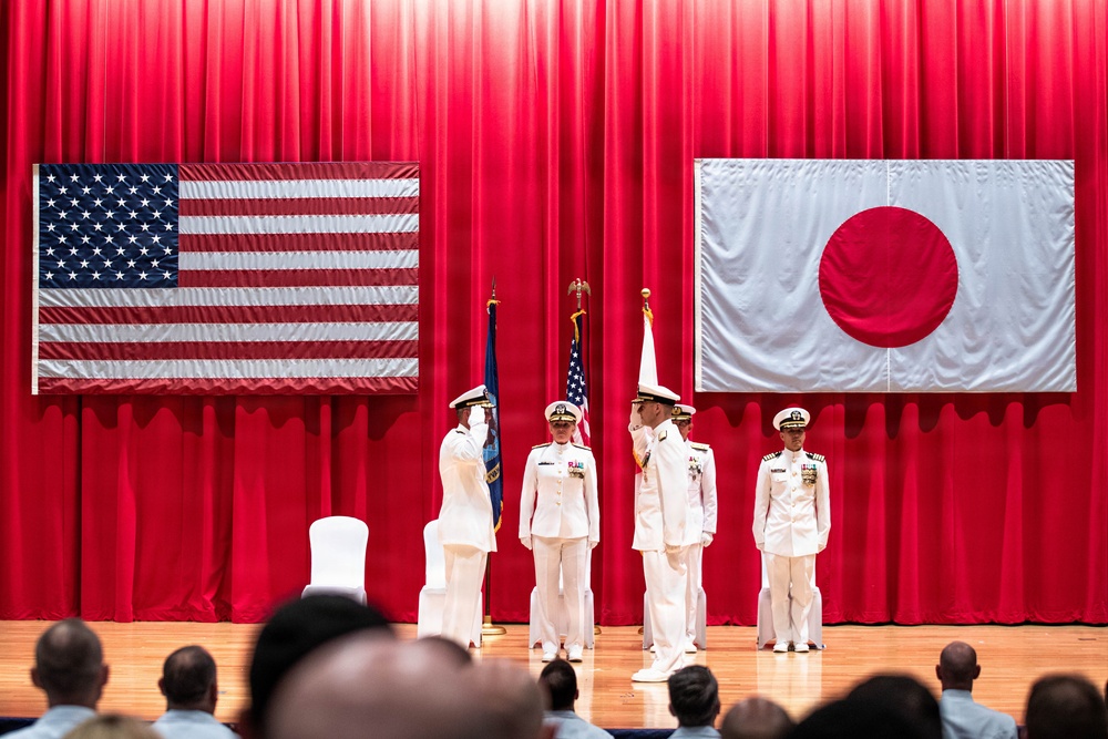 A formal military ceremony is taking place on a stage with red curtains, featuring personnel in white dress uniforms. Two large flags — the United States flag and the Japanese flag — are prominently displayed on the back wall, indicating a joint US-Japan military event. Several uniformed officers are positioned on stage, with two appearing to exchange salutes, suggesting a change of command or similar official ceremony.