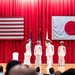 A formal military ceremony is taking place on a stage with red curtains, featuring personnel in white dress uniforms. Two large flags — the United States flag and the Japanese flag — are prominently displayed on the back wall, indicating a joint US-Japan military event. Several uniformed officers are positioned on stage, with two appearing to exchange salutes, suggesting a change of command or similar official ceremony.