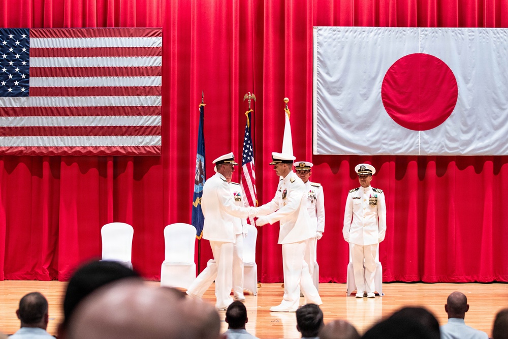 A formal military ceremony is taking place on a stage decorated with the flags of the United States and Japan, suggesting a joint US-Japan military event. Two naval officers in white dress uniforms are shaking hands at center stage, likely during a change of command or official recognition ceremony. Additional officers stand at attention in the background while an audience observes from the foreground.