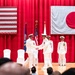 A formal military ceremony is taking place on a stage decorated with the flags of the United States and Japan, suggesting a joint US-Japan military event. Two naval officers in white dress uniforms are shaking hands at center stage, likely during a change of command or official recognition ceremony. Additional officers stand at attention in the background while an audience observes from the foreground.