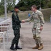 Two military personnel are shaking hands outdoors on a paved walkway near a building. One individual wears a dark green camouflage uniform with a black beret, consistent with Southeast Asian military dress, while the other wears a US Army MultiCam (OCP) uniform with visible 'US ARMY' tape. The interaction appears to be a formal greeting or diplomatic meeting between allied military officers.