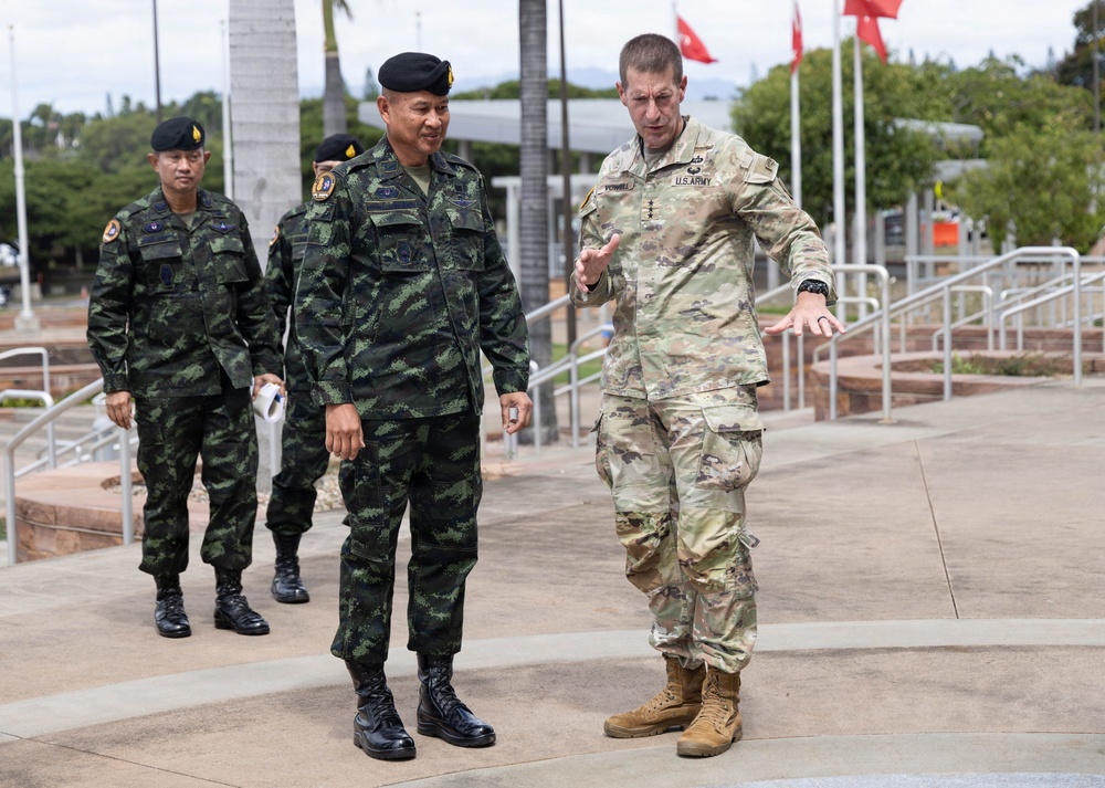 A U.S. Army officer in MultiCam/OCP uniform is walking and conversing with a senior officer wearing Thai Royal Army camouflage uniform and black beret. Two additional Thai military personnel follow behind them in the background. The setting appears to be an outdoor military installation, likely in Hawaii based on the tropical vegetation and architectural style.