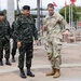 A U.S. Army officer in MultiCam/OCP uniform is walking and conversing with a senior officer wearing Thai Royal Army camouflage uniform and black beret. Two additional Thai military personnel follow behind them in the background. The setting appears to be an outdoor military installation, likely in Hawaii based on the tropical vegetation and architectural style.