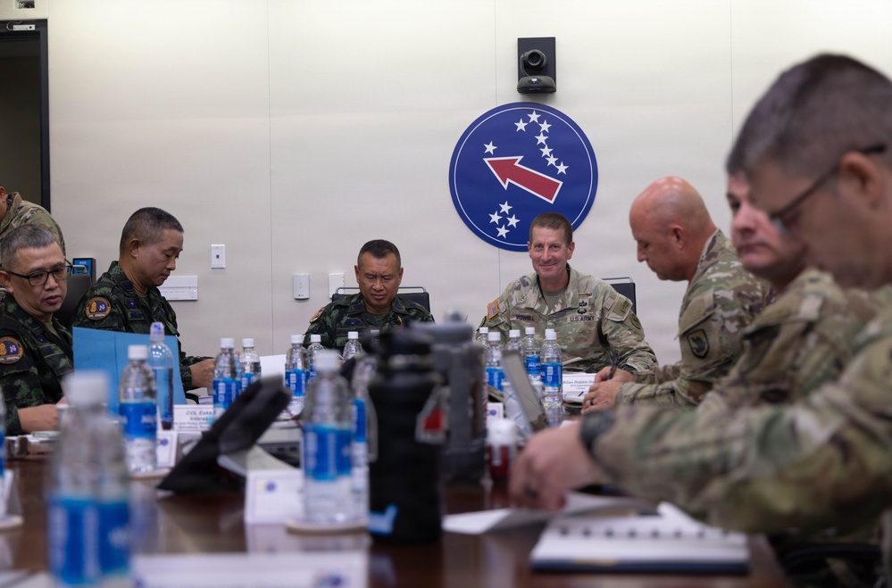 A military meeting or briefing is taking place around a conference table, involving both U.S. Army personnel in OCP (Operational Camouflage Pattern) uniforms and foreign military officers in different camouflage uniforms. A blue circular insignia with stars and a red arrow — consistent with U.S. Army Pacific (USARPAC) — is prominently displayed on the wall behind the group. The setting suggests a bilateral or multinational military coordination meeting.