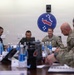 A military meeting or briefing is taking place around a conference table, involving both U.S. Army personnel in OCP (Operational Camouflage Pattern) uniforms and foreign military officers in different camouflage uniforms. A blue circular insignia with stars and a red arrow — consistent with U.S. Army Pacific (USARPAC) — is prominently displayed on the wall behind the group. The setting suggests a bilateral or multinational military coordination meeting.