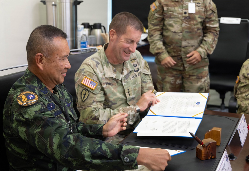 Two military officers — one in Thai Royal Army camouflage uniform and one in U.S. Army OCP (Operational Camouflage Pattern) uniform — are seated at a table reviewing or signing official documents in a formal meeting setting. The U.S. officer displays two-star general rank insignia and a 25th Infantry Division patch. A third soldier in U.S. Army OCP uniform stands in the background observing the proceedings.