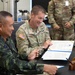 Two military officers — one in Thai Royal Army camouflage uniform and one in U.S. Army OCP (Operational Camouflage Pattern) uniform — are seated at a table reviewing or signing official documents in a formal meeting setting. The U.S. officer displays two-star general rank insignia and a 25th Infantry Division patch. A third soldier in U.S. Army OCP uniform stands in the background observing the proceedings.