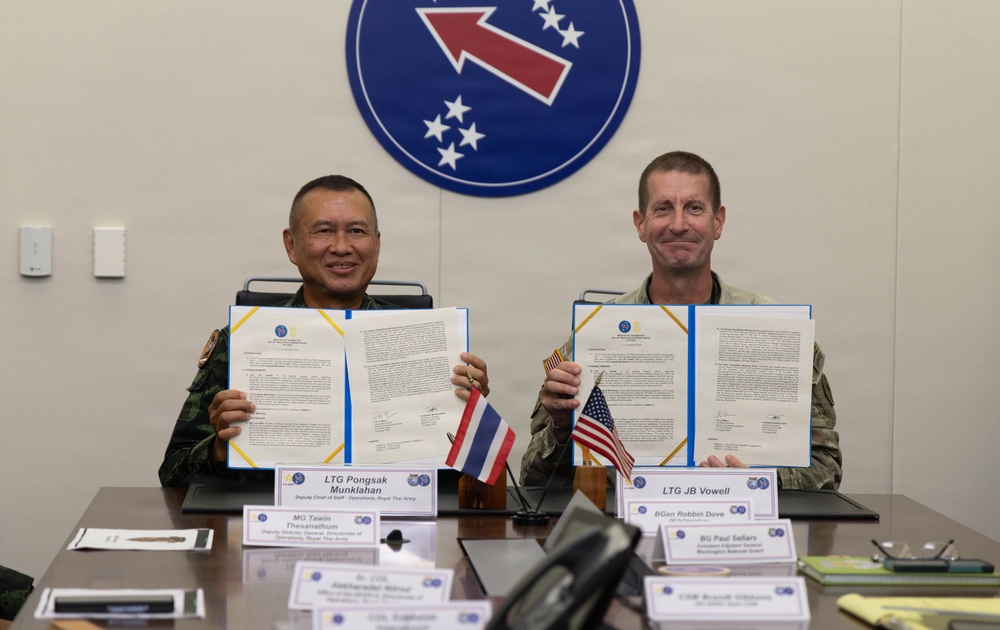 Two military officers, one from the Royal Thai Army and one from the U.S. Army, are seated at a conference table holding up signed documents in blue folders. Name placards identify them as LTG Pongsak Munklahan (Deputy Chief of Staff Operations, Royal Thai Army) and LTG JB Vowell. The setting appears to be a formal bilateral military agreement signing ceremony, with Thai and American flags visible on the table and a military unit insignia (red arrow with stars) displayed on the wall behind them.