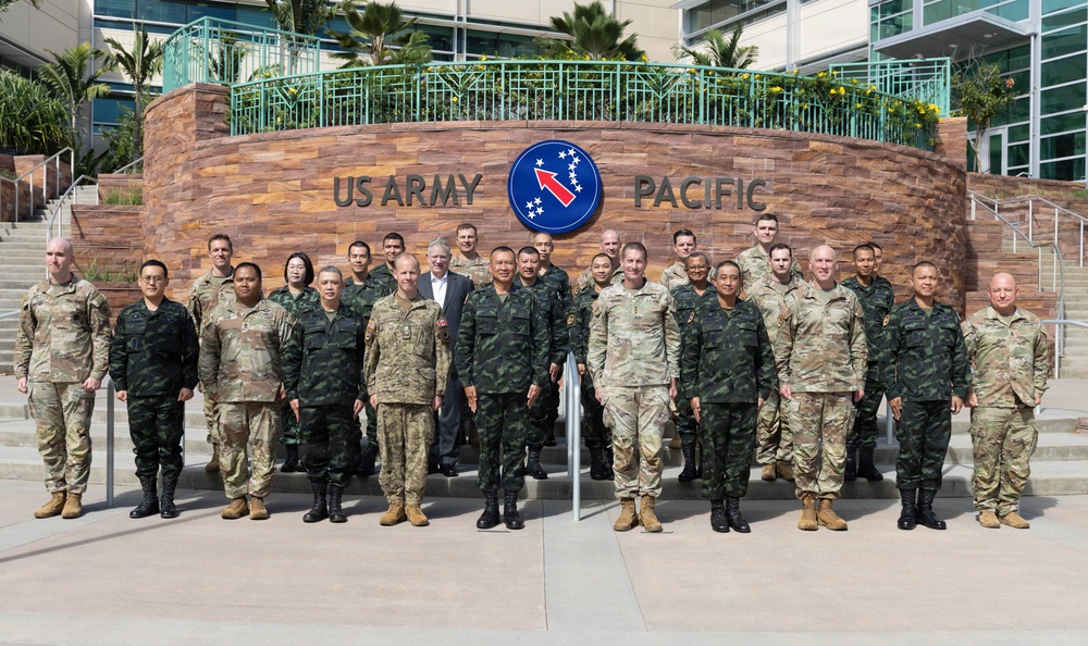 A group of approximately 25-30 military personnel pose for a formal group photograph in front of the US Army Pacific headquarters sign. The group includes personnel wearing multiple different camouflage uniform patterns, suggesting a multinational military gathering or delegation visit. One civilian in a white shirt is also present among the uniformed personnel.
