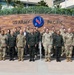 A group of approximately 25-30 military personnel pose for a formal group photograph in front of the US Army Pacific headquarters sign. The group includes personnel wearing multiple different camouflage uniform patterns, suggesting a multinational military gathering or delegation visit. One civilian in a white shirt is also present among the uniformed personnel.