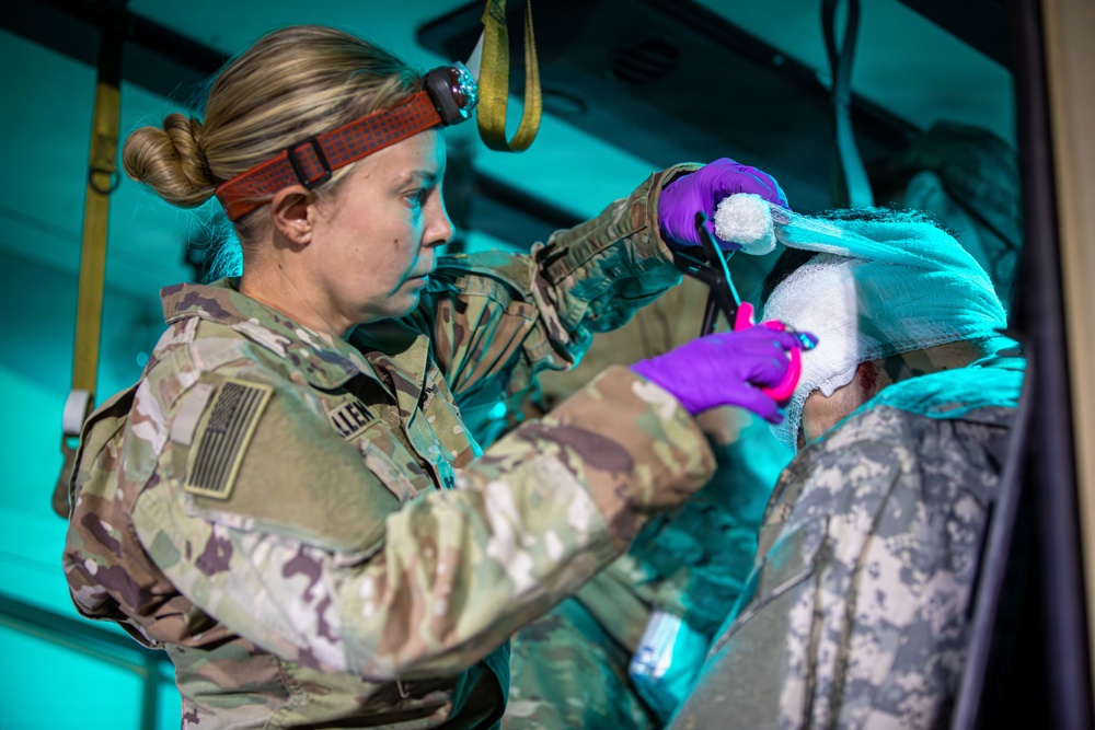 A U.S. Army medic wearing OCP (Operational Camouflage Pattern) uniform and a red headlamp is providing medical treatment to a patient whose head is wrapped in bandages. The medic, whose name tape reads 'ALLEN', is wearing purple nitrile gloves and appears to be applying or adjusting wound dressings to the patient's head. The scene appears to take place inside a military medical vehicle or tent under teal/blue ambient lighting.