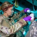 A U.S. Army medic wearing OCP (Operational Camouflage Pattern) uniform and a red headlamp is providing medical treatment to a patient whose head is wrapped in bandages. The medic, whose name tape reads 'ALLEN', is wearing purple nitrile gloves and appears to be applying or adjusting wound dressings to the patient's head. The scene appears to take place inside a military medical vehicle or tent under teal/blue ambient lighting.