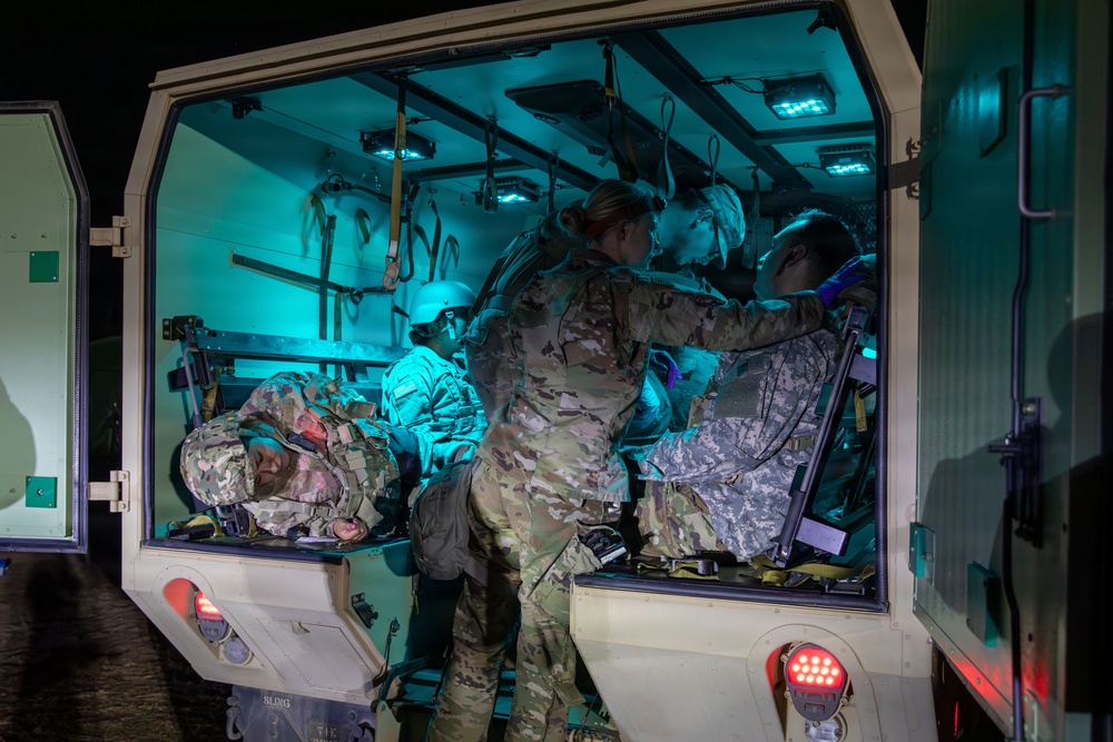 Military personnel in camouflage uniforms are conducting a medical evacuation or casualty care exercise inside what appears to be a military ambulance or medical transport vehicle at night. At least two individuals appear to be acting as patients on litters/stretchers while a standing medic attends to them. The interior is illuminated by teal/cyan-colored lighting, suggesting a tactical night operations scenario.