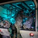 Military personnel in camouflage uniforms are conducting a medical evacuation or casualty care exercise inside what appears to be a military ambulance or medical transport vehicle at night. At least two individuals appear to be acting as patients on litters/stretchers while a standing medic attends to them. The interior is illuminated by teal/cyan-colored lighting, suggesting a tactical night operations scenario.