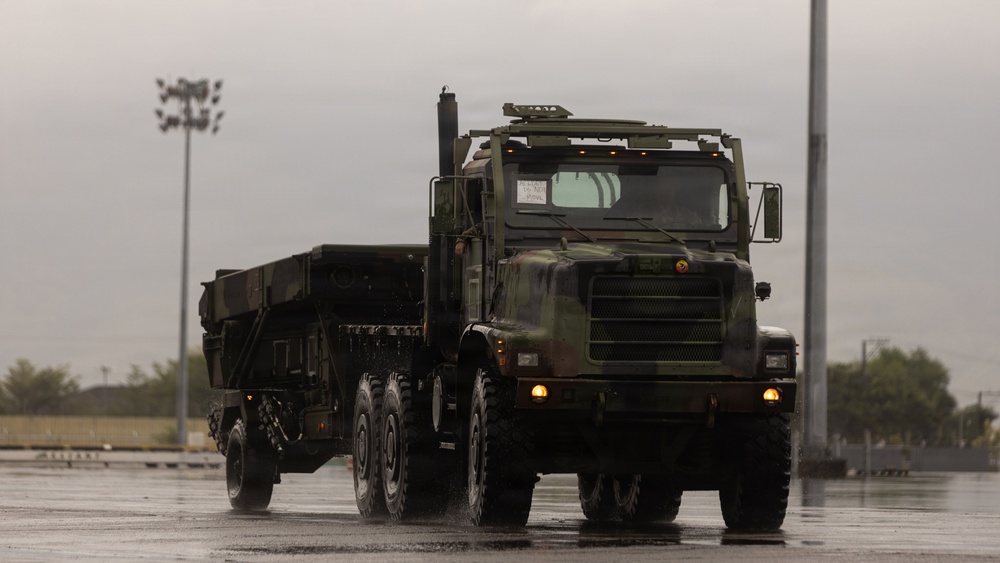 A large military tactical truck in camouflage paint scheme is driving on a wet surface during rainy conditions. The vehicle appears to be a heavy-duty multi-axle military transport truck, likely a variant of the Oshkosh HEMTT or similar platform, pulling a trailer. The overcast, wet weather conditions and reflective pavement suggest active operations or a military installation exercise.