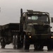 A large military tactical truck in camouflage paint scheme is driving on a wet surface during rainy conditions. The vehicle appears to be a heavy-duty multi-axle military transport truck, likely a variant of the Oshkosh HEMTT or similar platform, pulling a trailer. The overcast, wet weather conditions and reflective pavement suggest active operations or a military installation exercise.