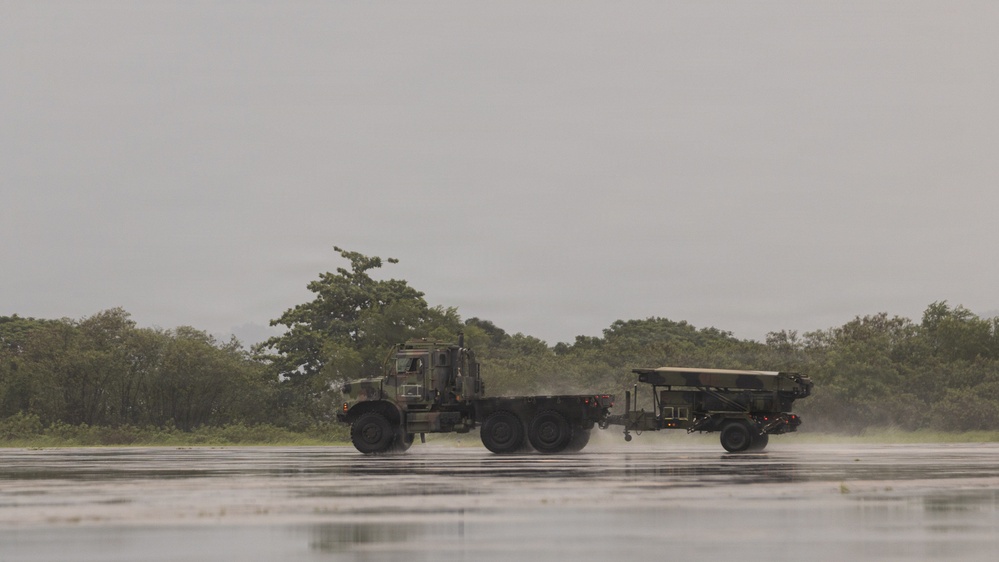 A military truck, appearing to be a medium tactical vehicle (MTV) in olive drab/camouflage paint, is towing a trailer-mounted weapons system across a wet tarmac or airstrip surface. The scene appears to be taken during or after rainfall, with water reflections visible on the ground surface. The setting includes dense tropical or subtropical vegetation in the background under an overcast sky.