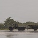 A military truck, appearing to be a medium tactical vehicle (MTV) in olive drab/camouflage paint, is towing a trailer-mounted weapons system across a wet tarmac or airstrip surface. The scene appears to be taken during or after rainfall, with water reflections visible on the ground surface. The setting includes dense tropical or subtropical vegetation in the background under an overcast sky.