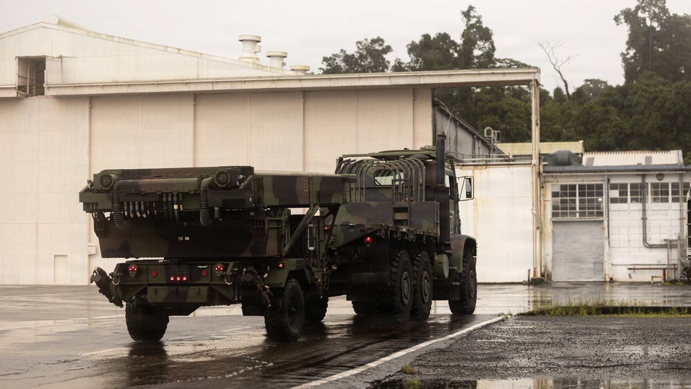 The image shows two military vehicles in camouflage paint scheme parked on a wet tarmac or paved area near a large white industrial or military building. The lead vehicle appears to be an M270 Multiple Launch Rocket System (MLRS) or similar rocket artillery platform mounted on a tracked/wheeled chassis. The scene appears to be taken during or after rainfall at what looks like a military installation.