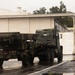 The image shows two military vehicles in camouflage paint scheme parked on a wet tarmac or paved area near a large white industrial or military building. The lead vehicle appears to be an M270 Multiple Launch Rocket System (MLRS) or similar rocket artillery platform mounted on a tracked/wheeled chassis. The scene appears to be taken during or after rainfall at what looks like a military installation.