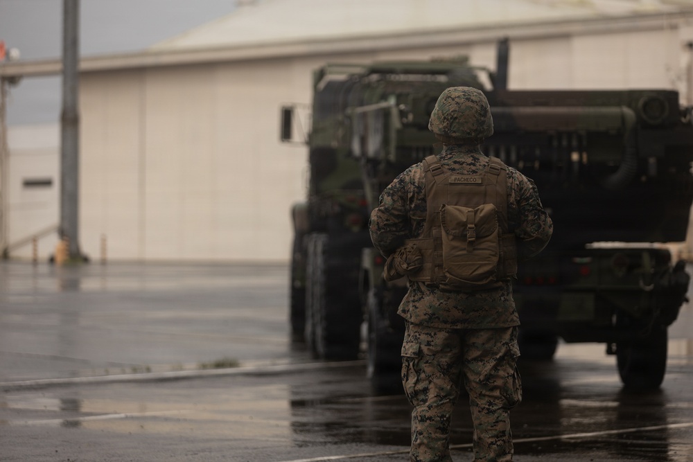 A U.S. Marine, identified by the name tape 'PACHECO' on their body armor, stands on a wet tarmac or paved surface facing a large military tactical vehicle. The Marine is wearing full combat gear including MARPAT camouflage uniform, helmet, and a coyote-tan plate carrier with an attached pouch. The overcast, rainy conditions and the presence of a large building in the background suggest a military installation or airfield.