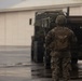 A U.S. Marine, identified by the name tape 'PACHECO' on their body armor, stands on a wet tarmac or paved surface facing a large military tactical vehicle. The Marine is wearing full combat gear including MARPAT camouflage uniform, helmet, and a coyote-tan plate carrier with an attached pouch. The overcast, rainy conditions and the presence of a large building in the background suggest a military installation or airfield.