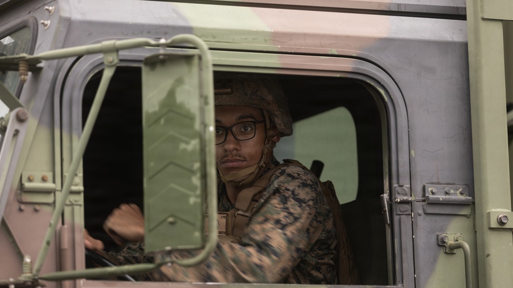 A military service member wearing digital camouflage uniform and a combat helmet is seated in the cab of a military tactical vehicle. The individual is wearing black-framed glasses and appears to be in the driver's seat, looking toward the camera. The vehicle appears to be a standard military utility truck with olive drab/sage green coloring.