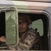 A military service member wearing digital camouflage uniform and a combat helmet is seated in the cab of a military tactical vehicle. The individual is wearing black-framed glasses and appears to be in the driver's seat, looking toward the camera. The vehicle appears to be a standard military utility truck with olive drab/sage green coloring.