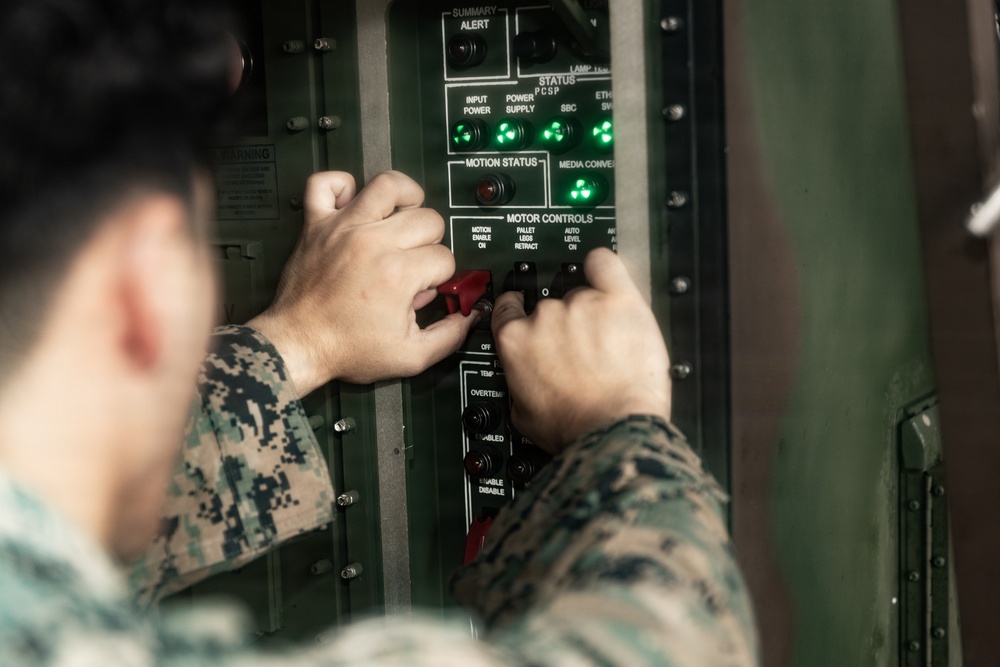 A U.S. Marine in digital camouflage (MARPAT) uniform is operating a military control panel with both hands. The panel contains status indicator lights, motion status displays, and motor control switches, several of which appear to be red-handled toggle switches. The green indicator lights on the STATUS section of the panel are illuminated, suggesting the system is powered and operational.