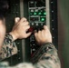 A U.S. Marine in digital camouflage (MARPAT) uniform is operating a military control panel with both hands. The panel contains status indicator lights, motion status displays, and motor control switches, several of which appear to be red-handled toggle switches. The green indicator lights on the STATUS section of the panel are illuminated, suggesting the system is powered and operational.