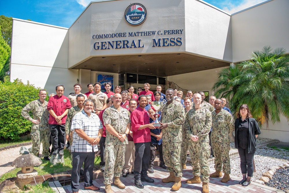 A group of military personnel in U.S. Navy camouflage uniforms and civilian staff pose for a group photo in front of the Commodore Matthew C. Perry General Mess facility, which bears the CFAY (Commander Fleet Activities Yokosuka) emblem. Two individuals in the center are presenting or holding a trophy, suggesting an award or recognition ceremony. The setting appears to be a U.S. Navy installation, likely in Japan based on the CFAY insignia and the mix of American military and Japanese civilian staff.