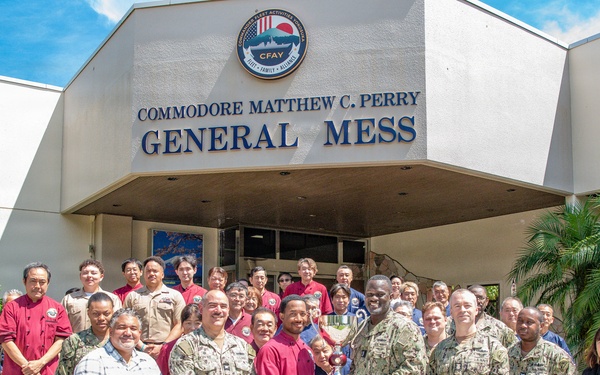 A group of military personnel in U.S. Navy camouflage uniforms and civilian staff pose for a group photo in front of the Commodore Matthew C. Perry General Mess facility, which bears the CFAY (Commander Fleet Activities Yokosuka) emblem. Two individuals in the center are presenting or holding a trophy, suggesting an award or recognition ceremony. The setting appears to be a U.S. Navy installation, likely in Japan based on the CFAY insignia and the mix of American military and Japanese civilian staff.