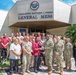 A group of military personnel in U.S. Navy camouflage uniforms and civilian staff pose for a group photo in front of the Commodore Matthew C. Perry General Mess facility, which bears the CFAY (Commander Fleet Activities Yokosuka) emblem. Two individuals in the center are presenting or holding a trophy, suggesting an award or recognition ceremony. The setting appears to be a U.S. Navy installation, likely in Japan based on the CFAY insignia and the mix of American military and Japanese civilian staff.