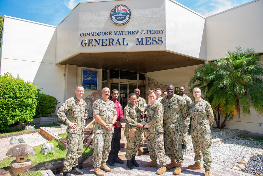 A group of approximately 10-11 U.S. Navy personnel in NWU (Navy Working Uniform) camouflage are posing for a group photo in front of the 'Commodore Matthew C. Perry General Mess' building at Commander Fleet Activities Yokosuka (CFAY), Japan. Two individuals in the center are holding a trophy or award, suggesting a recognition or award ceremony. The group includes both uniformed military members and at least one civilian in a red shirt.