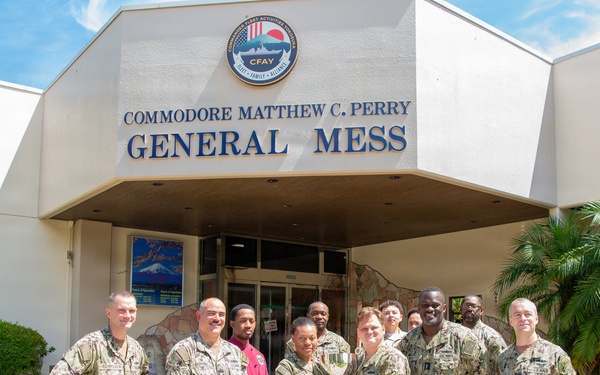 A group of approximately 10-11 U.S. Navy personnel in NWU (Navy Working Uniform) camouflage are posing for a group photo in front of the 'Commodore Matthew C. Perry General Mess' building at Commander Fleet Activities Yokosuka (CFAY), Japan. Two individuals in the center are holding a trophy or award, suggesting a recognition or award ceremony. The group includes both uniformed military members and at least one civilian in a red shirt.