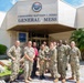 A group of approximately 10-11 U.S. Navy personnel in NWU (Navy Working Uniform) camouflage are posing for a group photo in front of the 'Commodore Matthew C. Perry General Mess' building at Commander Fleet Activities Yokosuka (CFAY), Japan. Two individuals in the center are holding a trophy or award, suggesting a recognition or award ceremony. The group includes both uniformed military members and at least one civilian in a red shirt.