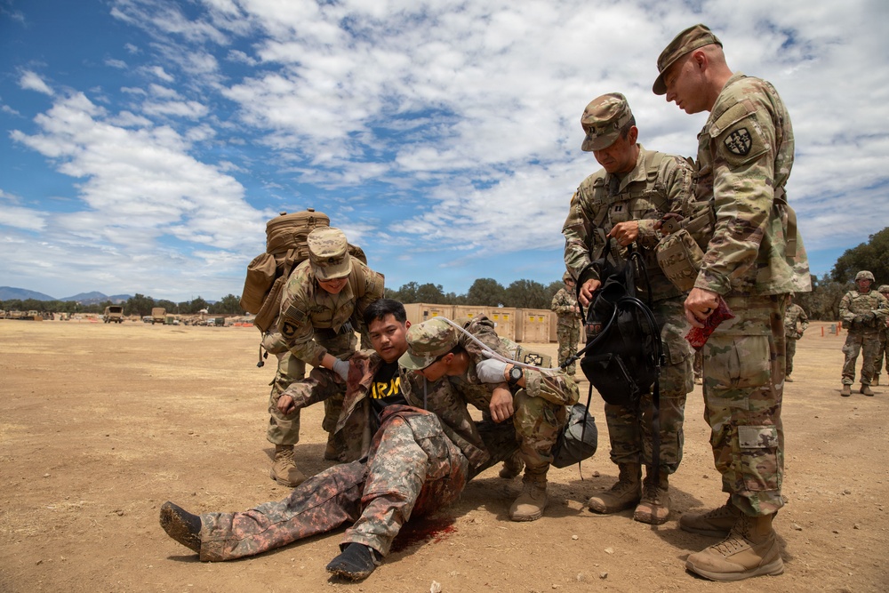 A military medical training exercise is taking place on an outdoor dirt field. Several soldiers in U.S. Army OCP (Operational Camouflage Pattern) uniforms are attending to a simulated casualty lying on the ground with mock injuries. The scene depicts a Combat Lifesaver or TCCC (Tactical Combat Casualty Care) training scenario.