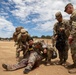 A military medical training exercise is taking place on an outdoor dirt field. Several soldiers in U.S. Army OCP (Operational Camouflage Pattern) uniforms are attending to a simulated casualty lying on the ground with mock injuries. The scene depicts a Combat Lifesaver or TCCC (Tactical Combat Casualty Care) training scenario.