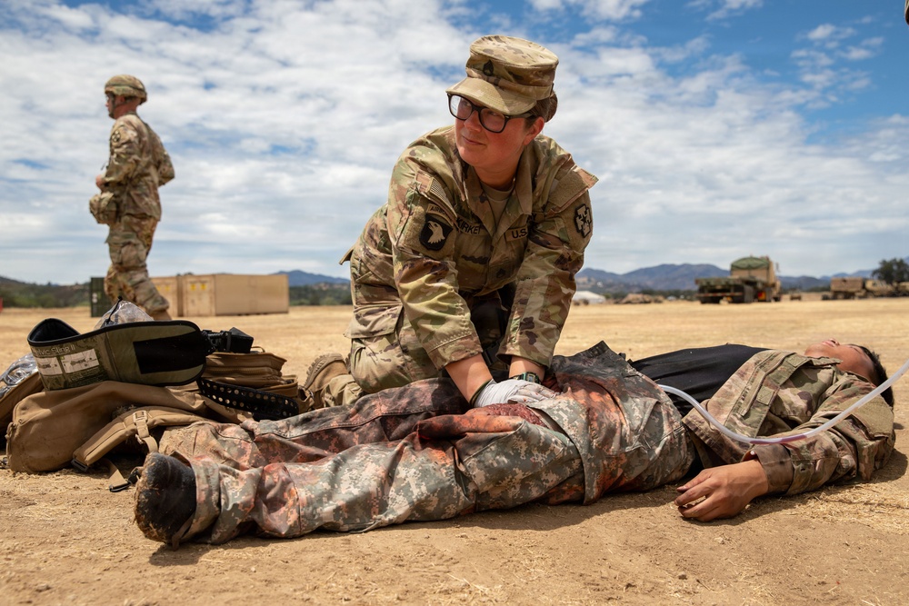 A U.S. Army soldier in OCP (Operational Camouflage Pattern) uniform is performing combat casualty care on a simulated wounded soldier lying on the ground during what appears to be a field training exercise. The 'casualty' is wearing older ACU (Army Combat Uniform) pattern and has simulated blood/wounds on the torso, with a medical tube visible near the neck area. A second soldier stands in the background near military equipment and shipping containers.