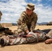 A U.S. Army soldier in OCP (Operational Camouflage Pattern) uniform is performing combat casualty care on a simulated wounded soldier lying on the ground during what appears to be a field training exercise. The 'casualty' is wearing older ACU (Army Combat Uniform) pattern and has simulated blood/wounds on the torso, with a medical tube visible near the neck area. A second soldier stands in the background near military equipment and shipping containers.