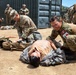 A military medical training exercise is underway in an outdoor, arid environment. Soldiers in camouflage uniforms, including one with a visible Canadian Armed Forces flag patch and red cross medical insignia, are performing casualty care on a simulated wounded soldier lying on the ground. Multiple personnel are engaged in similar activities across the scene, indicating a large-scale combat casualty care drill.