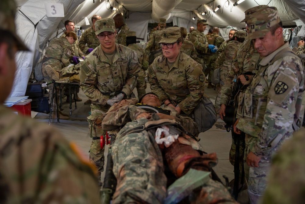 U.S. Army medical personnel are conducting what appears to be a field medical training exercise inside a military medical tent. Soldiers wearing OCP (Operational Camouflage Pattern) uniforms are treating a simulated casualty on a litter/stretcher. The setting is a large white military field hospital tent with multiple treatment stations visible in the background.
