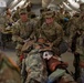 U.S. Army medical personnel are conducting what appears to be a field medical training exercise inside a military medical tent. Soldiers wearing OCP (Operational Camouflage Pattern) uniforms are treating a simulated casualty on a litter/stretcher. The setting is a large white military field hospital tent with multiple treatment stations visible in the background.