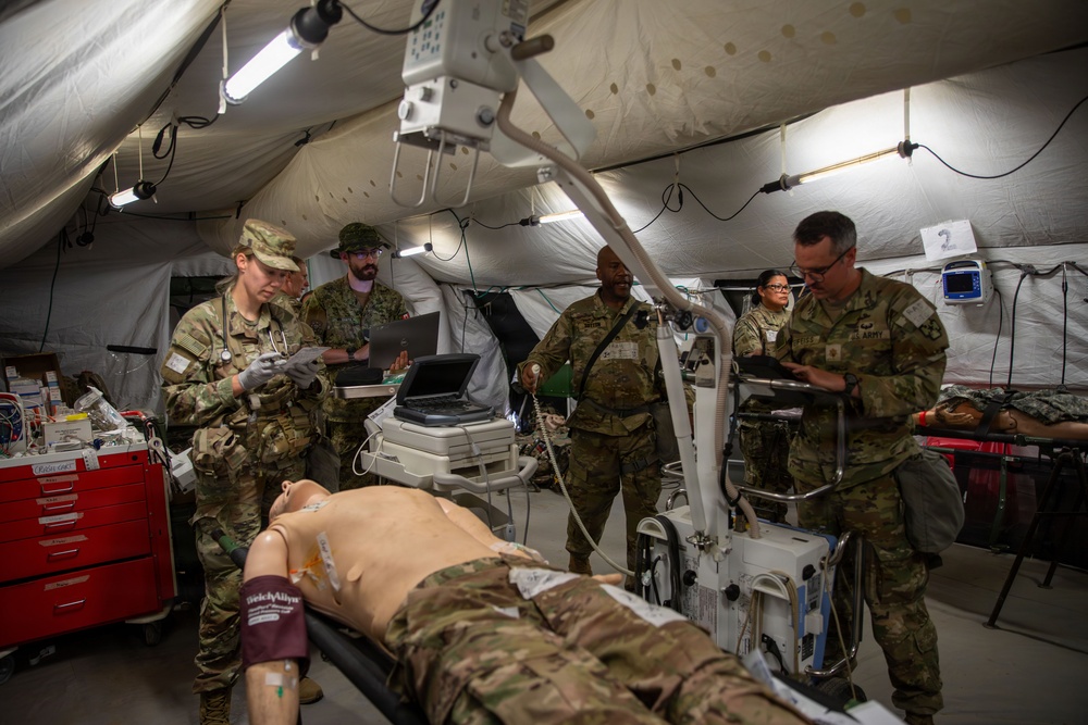 Military medical personnel are conducting a field hospital training exercise inside a large tent structure. A medical training mannequin is positioned on a gurney in the foreground while several soldiers in camouflage uniforms review data on laptops and tablets. The scene depicts a simulated trauma care scenario with medical equipment including monitors, an ultrasound machine, and a portable X-ray unit.