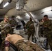 Military medical personnel are conducting a field hospital training exercise inside a large tent structure. A medical training mannequin is positioned on a gurney in the foreground while several soldiers in camouflage uniforms review data on laptops and tablets. The scene depicts a simulated trauma care scenario with medical equipment including monitors, an ultrasound machine, and a portable X-ray unit.