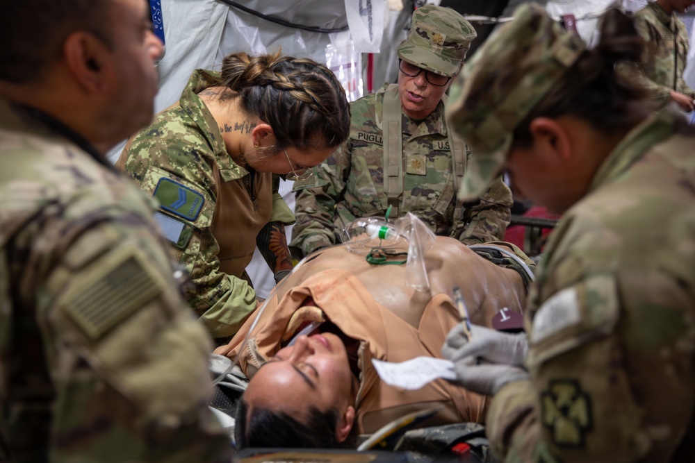 Military medical personnel in camouflage uniforms are attending to a patient lying on a stretcher, performing what appears to be a medical training exercise or field treatment scenario. Multiple soldiers surround the patient, who has a breathing mask/oxygen device on their chest. One soldier's name tape reading 'PUGLIA' is visible on their uniform.